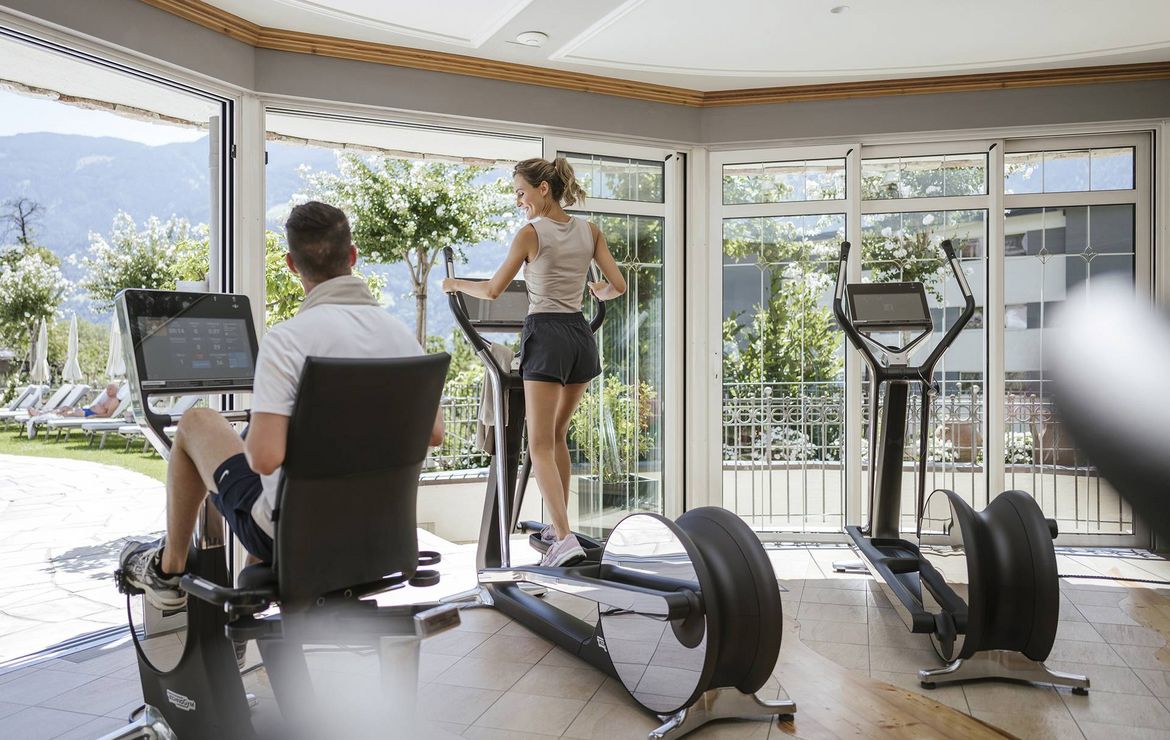 Eine Frau trainiert auf dem Crosstrainer, während ein Mann auf einem Ergometer im hellen Fitnessraum sitzt.