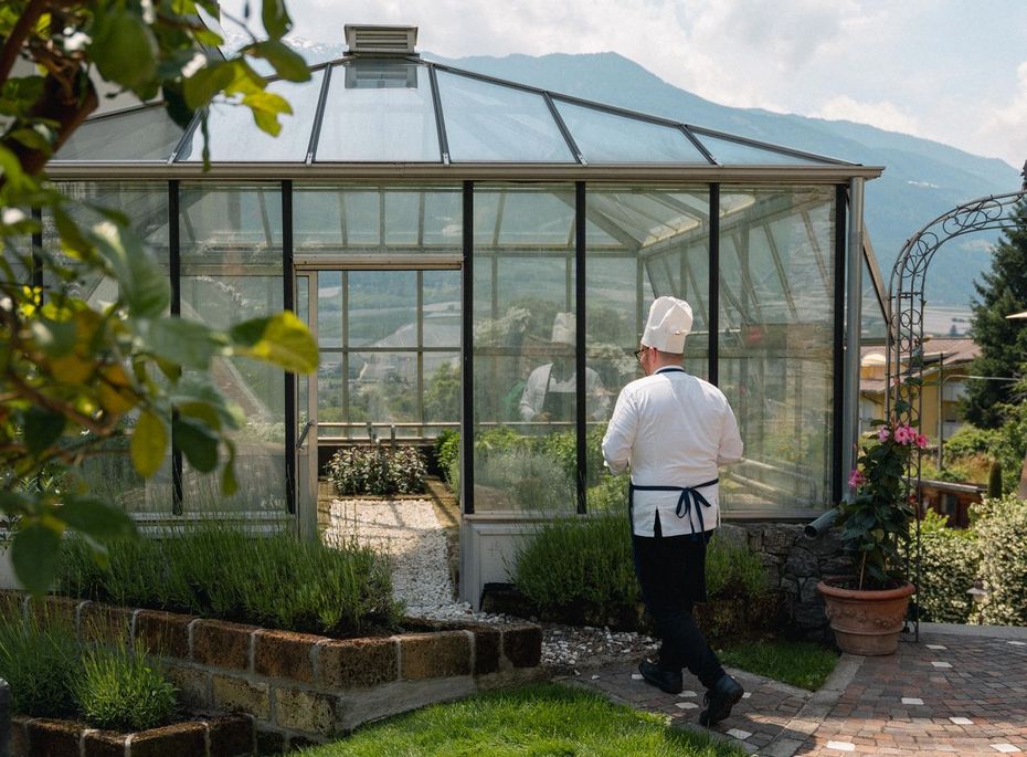 Ein Küchenchef geht auf einem Gartenweg auf ein Gewächshaus zu, im Hintergrund sind Berge zu sehen.