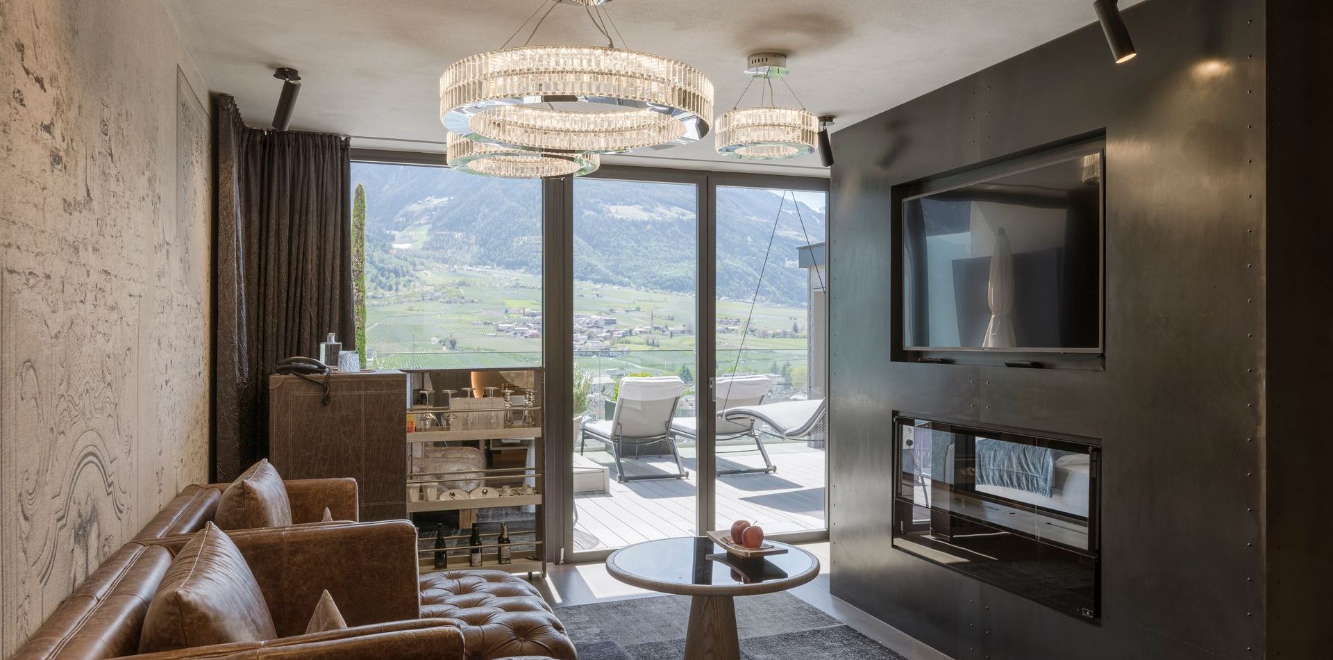 Ein Wohnbereich der Penthouse Suite mit Sofa, Kamin, TV-Wand und Blick auf den Balkon mit Bergpanorama.