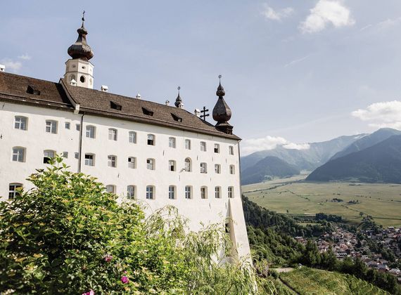Ein weißes Schloss mit Zwiebeltürmen liegt über dem Tal von Meran mit Bergen im Hintergrund.