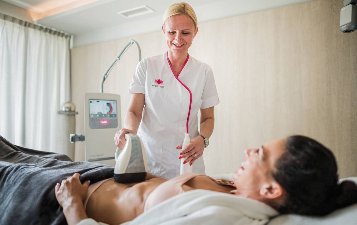 Eine Therapeutin führt eine Medical-Spa-Behandlung mit einem Handgerät am Bauch einer liegenden Frau durch.