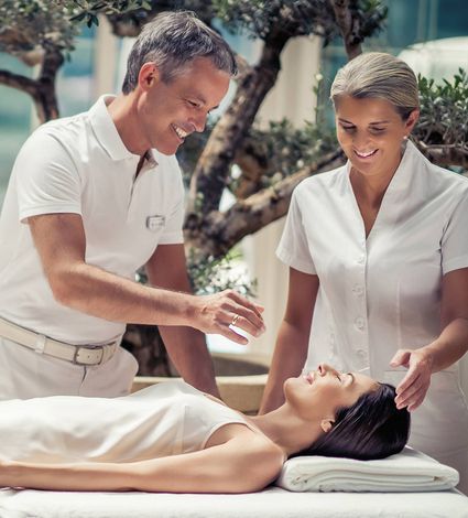 A therapist performs a spa treatment while a woman lies on a treatment bed in a calm wellness setting.