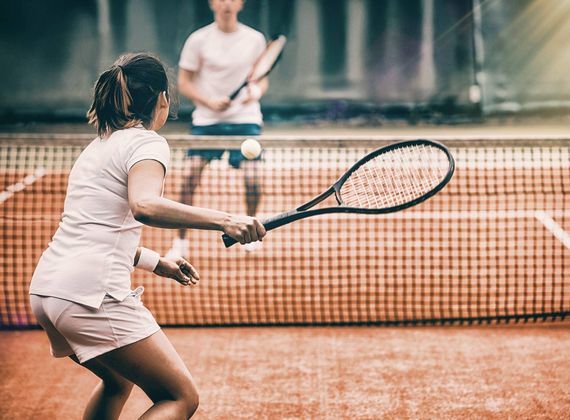 Eine Frau spielt auf dem Tennisplatz einen Ball an, während ihr Gegenüber am Netz bereitsteht.