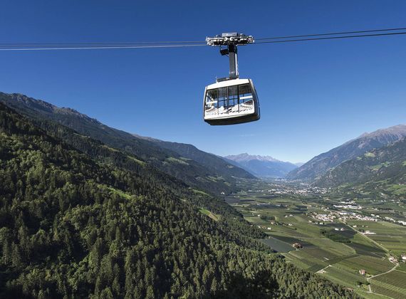 Eine Seilbahn schwebt über dem Tal mit Bergen, Wäldern und Feldern im Hintergrund.