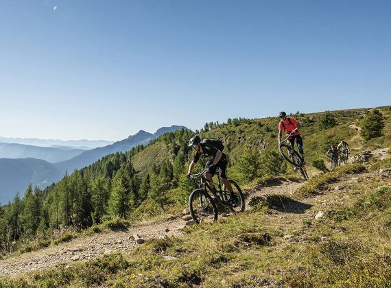 Drei Mountainbiker fahren auf einem schmalen Weg über einen sonnigen Bergrücken mit Blick ins Tal und auf die Berge.