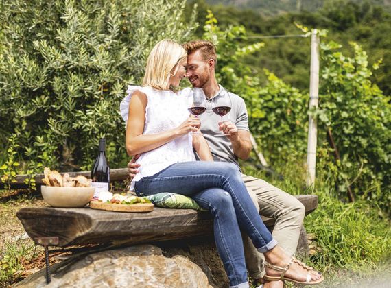 A couple sits outdoors in a garden and shares a wine tasting with glasses of red wine beside a small picnic table.