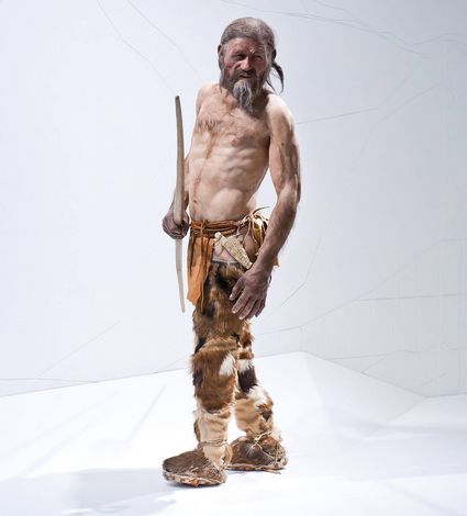 Ein Mann in historischer Fellkleidung steht auf hellem Hintergrund und hält einen langen Stock.