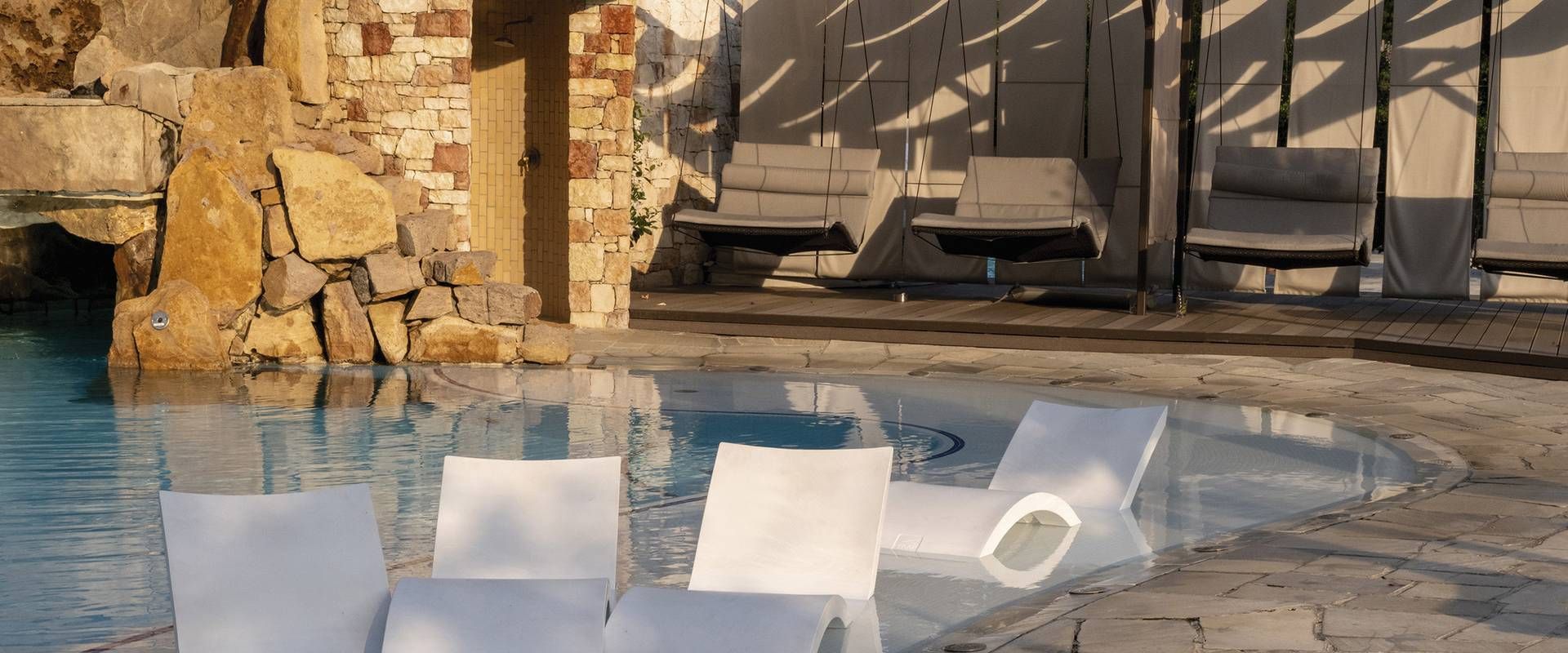 Lettini bianchi sono accanto a una tranquilla piscina spa interna con pareti in pietra e daybed sullo sfondo.
