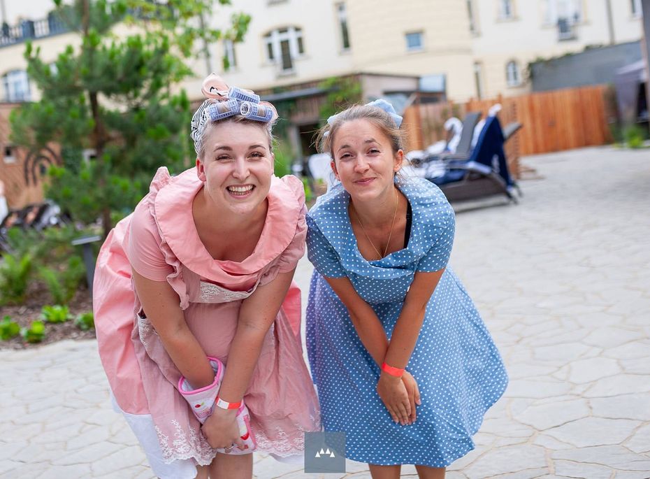 Zwei Frauen in farbigen Kleidern beugen sich lachend im Innenhof eines Hotels nach vorne.