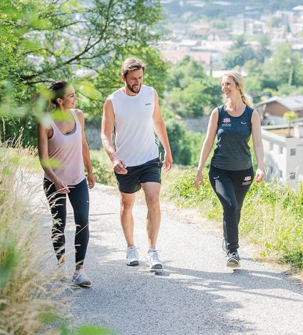 Drei Personen gehen auf einem Weg am Hang spazieren, mit Blick auf das Tal und die Ortschaft im Hintergrund.