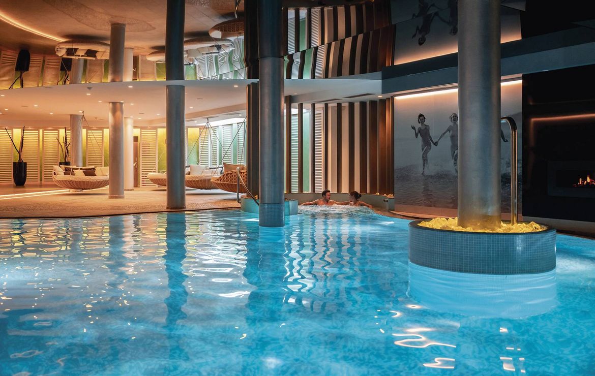 Una piscina spa interna con luci calde, lettini e colonne in un’area wellness moderna.