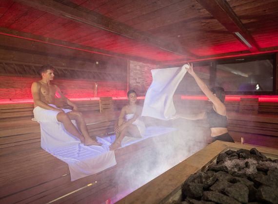 Drei Gäste erleben einen Saunaaufguss in der holzverkleideten Sauna mit aufsteigendem Dampf.