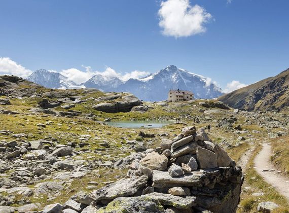 Eine Berghütte liegt in einer alpinen Landschaft mit Steinmännchen, Wanderweg und Bergen im Hintergrund.