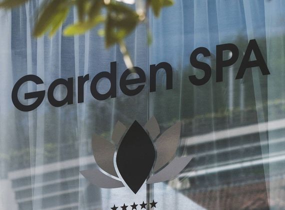 La scritta Garden Spa è visibile su una superficie in vetro con un logo a forma di loto e riflessi sfocati del verde sullo sfondo.