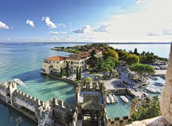 Blick auf die Halbinsel Sirmione mit Hafen, Uferpromenade und Booten im klaren Wasser des Gardasees.