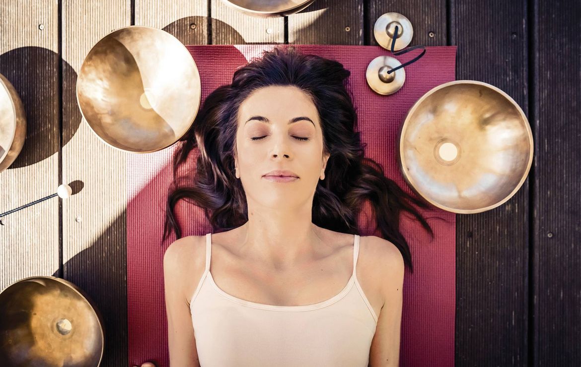 Eine Frau liegt entspannt auf einer Matte, umgeben von Klangschalen für eine Meditationsübung.