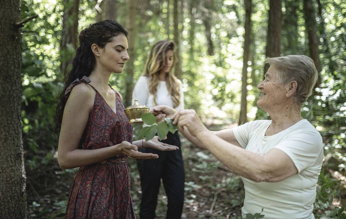 Eine Frau überreicht im Wald einer älteren Frau einen kleinen Teller mit Kräutern, während eine dritte Person im Hintergrund steht.