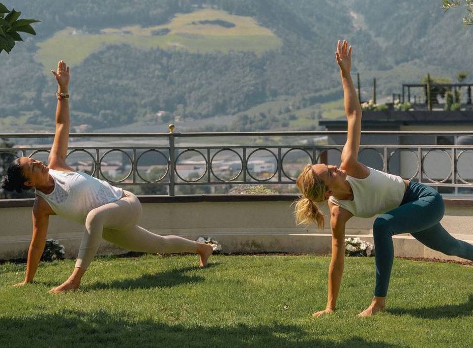Yoga auf der Terrasse im Preidlhof Zwei Frauen üben Yoga auf einer Rasenfläche mit Bergblick und Geländer im Hintergrund.