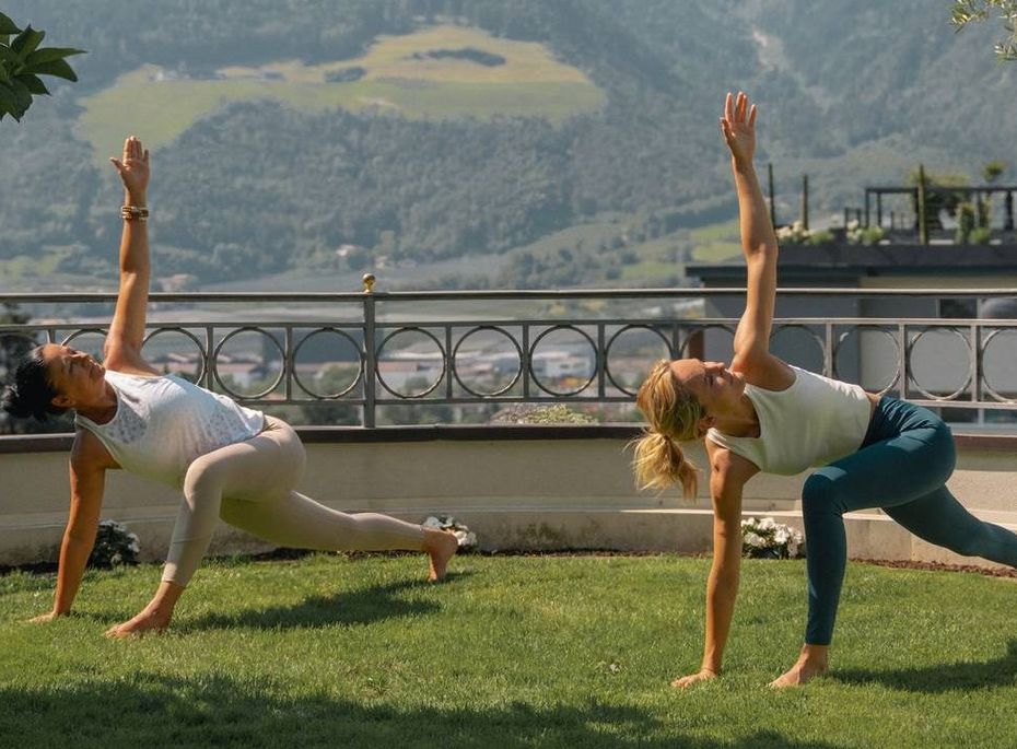 Yoga auf der Terrasse im Preidlhof Zwei Frauen üben Yoga auf einer Rasenfläche mit Bergblick und Geländer im Hintergrund.
