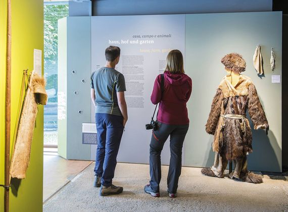 Zwei Personen betrachten eine Museumsausstellung mit historischen Gewändern und Texttafeln.