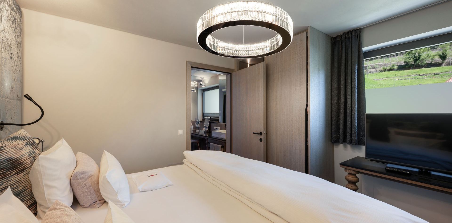 Ein modernes Schlafzimmer der Penthouse Suite mit Doppelbett, Kronleuchter, TV und Blick ins Grüne.