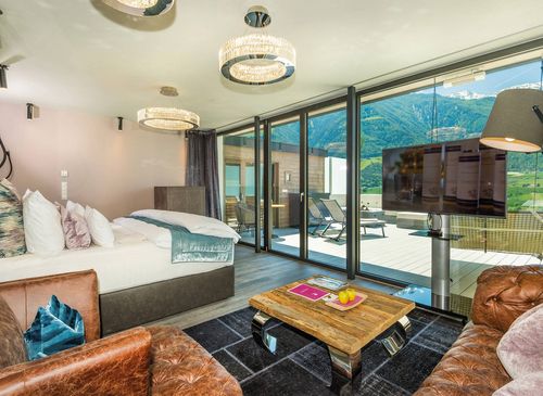 Penthouse Suite at Preidlhof with mountain view A penthouse suite with a bed, leather sofas, and a wall-mounted TV faces large glass doors to a balcony and mountains.