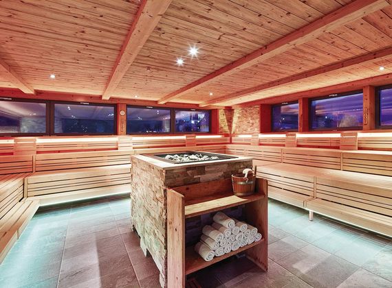 Eine helle Sauna mit Holzbank, Panoramafenstern und einem zentralen Ofen im Preidlhof.