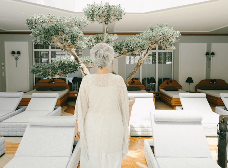 Ruheliegen im Wellnessbereich des Preidlhof Eine Frau steht vor weißen Ruheliegen im hellen Wellnessbereich mit Olivenbaum und großen Fenstern.