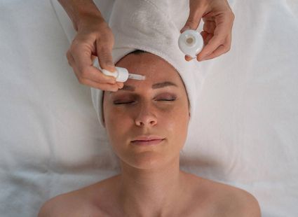 Eine Frau liegt bei einer Wellnessbehandlung mit Serum auf dem Gesicht und geschlossenen Augen auf der Liege.