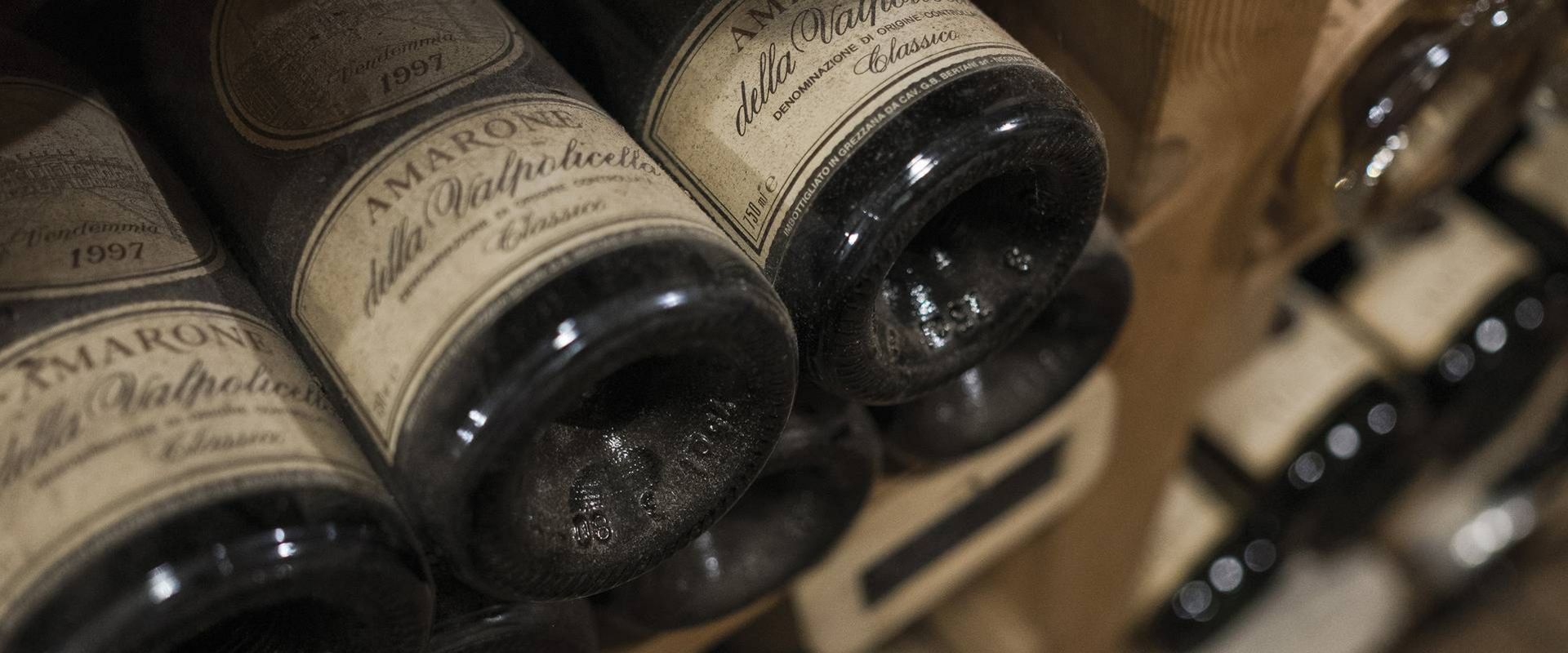 Più bottiglie di vino sono disposte in una rastrelliera in legno, con etichette e vetro impolverato in primo piano.
