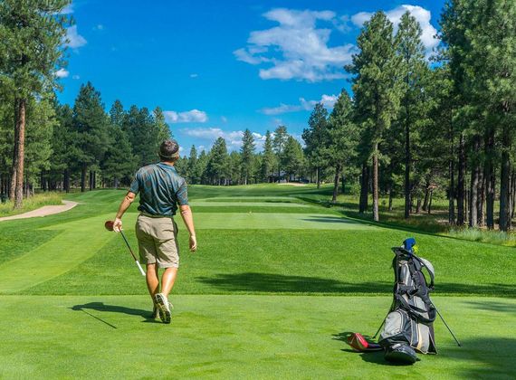 Ein Golfer geht über das Fairway auf einem Golfplatz, neben ihm steht ein Golfbag zwischen hohen Kiefern.