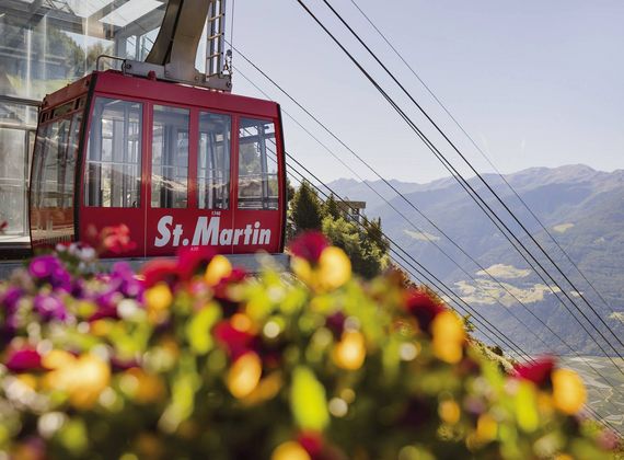 Eine rote Bergbahn namens St. Martin fährt an einer Bergstation vorbei, im Vordergrund sind unscharfe Blumen zu sehen.