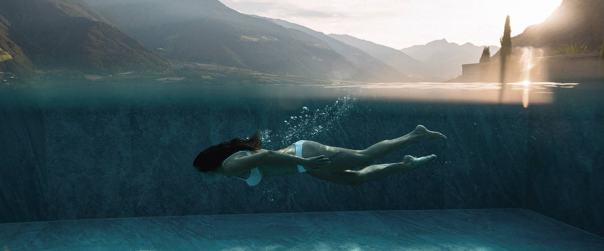 Eine Frau schwimmt in einem Infinity-Pool mit Blick auf die Berge und das helle Gegenlicht am Beckenrand.