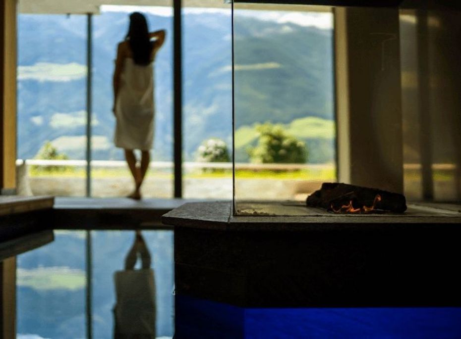 Eine Frau steht im Bademantel vor der Glasfront einer Sauna und blickt auf die Berge.