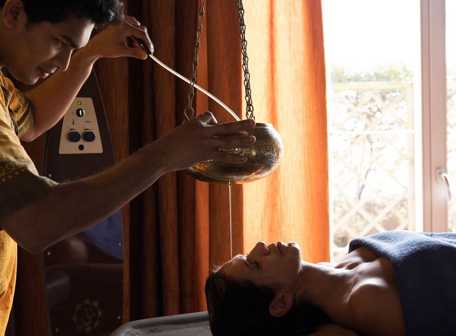 Ayurveda-Behandlung im Preidlhof Wellnesshotel Ein Behandler gießt warmes Öl aus einem Gefäß über die Stirn einer liegenden Frau bei einer Ayurveda-Behandlung.