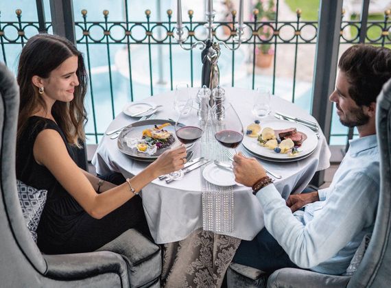 Una coppia è seduta a un tavolo con vino e piatti serviti accanto a una terrazza con piscina.