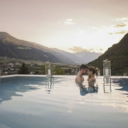 Ein Paar steht im Infinity-Pool und lehnt sich im Gegenlicht aneinander, dahinter öffnet sich der Blick auf das Tal und die Berge.