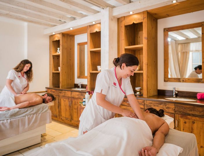 Zwei Therapeutinnen behandeln zwei Gäste gleichzeitig mit einer Wellnessmassage in einem hellen Spa-Raum.