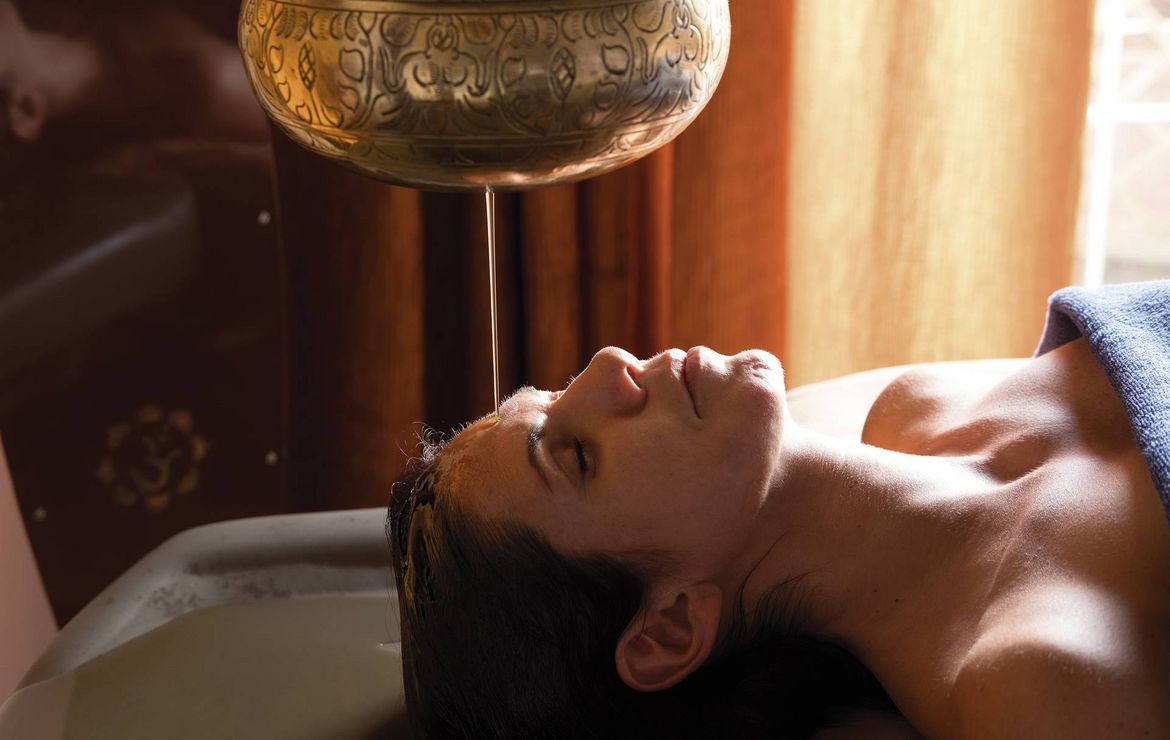 A woman lies back during an Ayurveda treatment as warm oil drips from a hanging vessel.