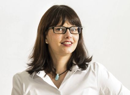 A woman with glasses and dark hair wears a white shirt against a plain light background.
