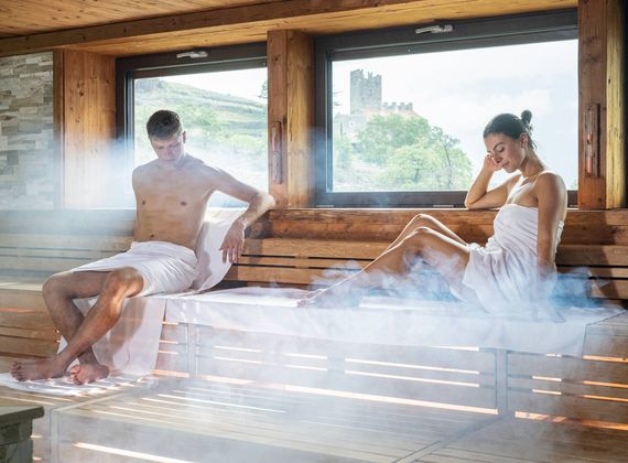 Ein Mann und eine Frau sitzen in einer holzverkleideten Sauna mit Dampf, durch das Fenster ist eine Burg zu sehen.