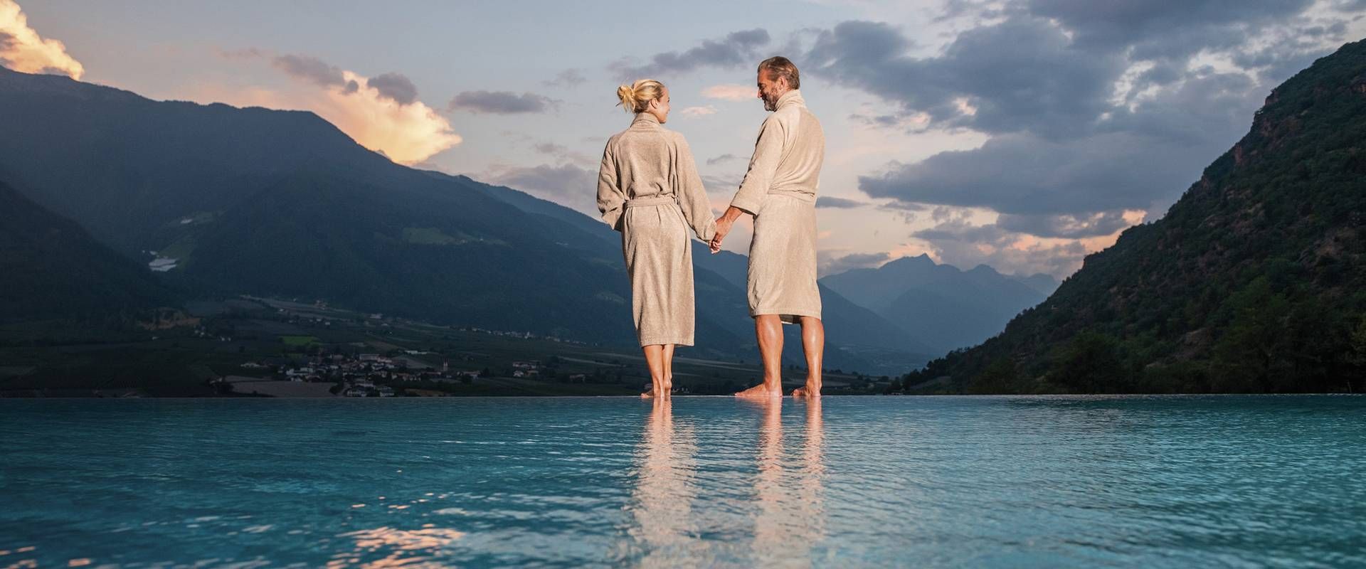 Ein Paar steht in einem Infinity-Pool und blickt über den See auf die Berge bei Sonnenuntergang.