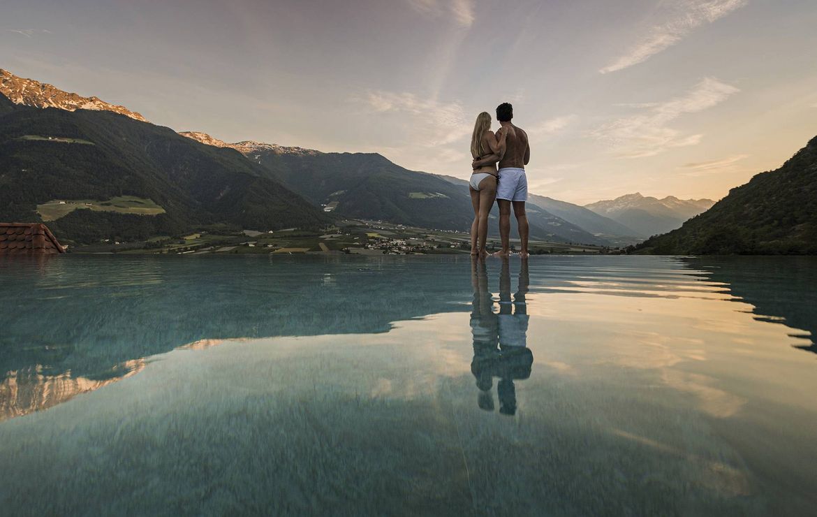 Ein Paar steht eng umschlungen am Rand des Infinity Pools mit Blick auf die Berge bei Sonnenuntergang.