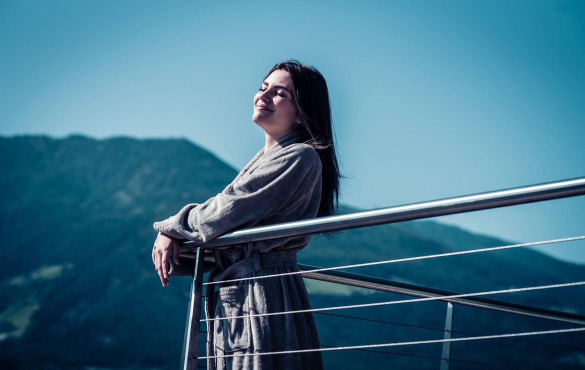 Una donna in accappatoio si rilassa su un balcone accanto a una ringhiera metallica con le montagne sullo sfondo.