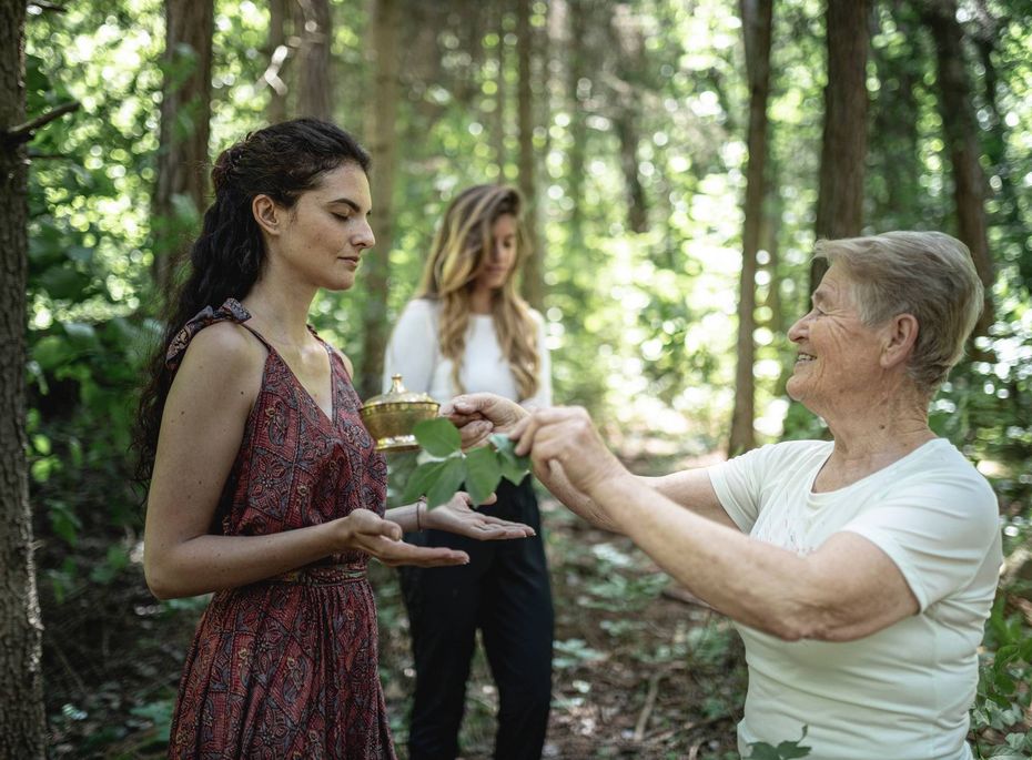 Waldritual im Wellnesshotel Preidlhof Eine ältere Frau reicht einer jungen Frau im Wald ein kleines Objekt, während eine dritte Person im Hintergrund steht.