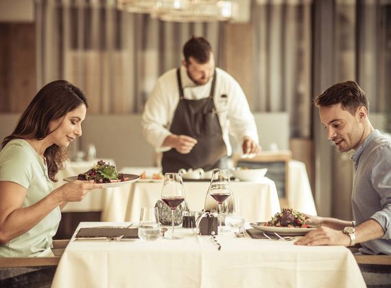 A couple enjoys a gourmet dinner while a chef serves a dish in the background.