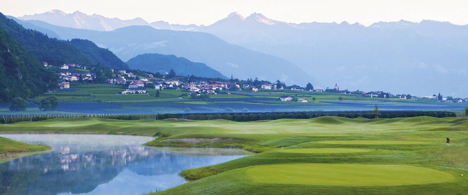 Ein weitläufiger Golfplatz im Meraner Land mit Wasserhindernis, grünen Fairways und Bergen im Hintergrund.
