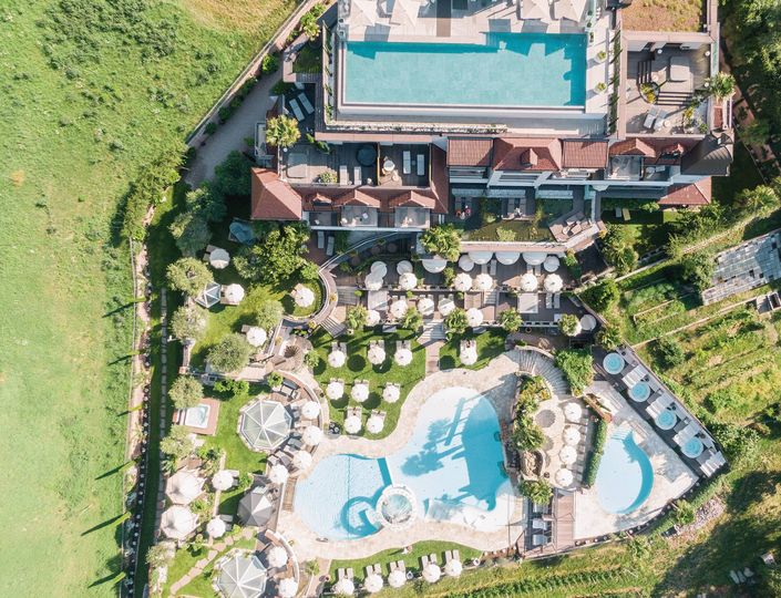 Blick von oben auf die Poollandschaft mit Infinity Pool, mehreren Liegeflächen und dem Hotelgebäude im Grünen.