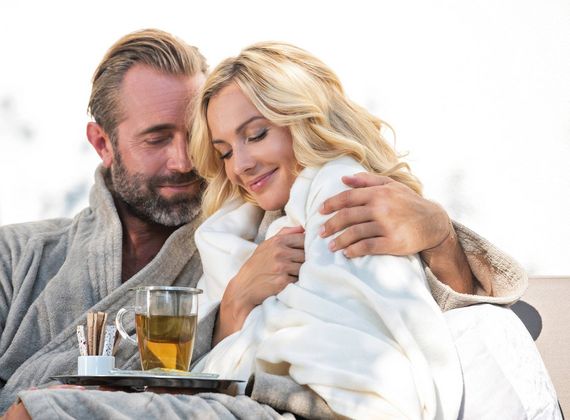 Ein Paar sitzt eng umschlungen mit Decken und einer Tasse Tee in einer entspannten Wellness-Szene.