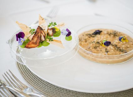 Ein fein angerichtetes Dessert mit Obst und Blüten steht neben einer Schale mit cremiger Speise auf einem weißen Teller.